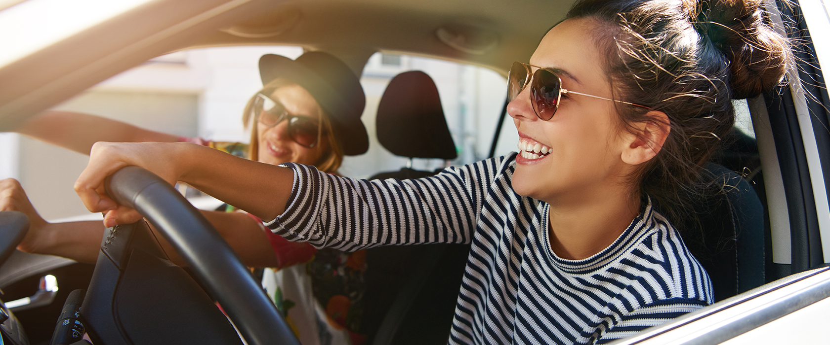 Zwei junge Frauen mit Sonnenbrillen fahren sorglos in einem Auto. Die Fenster sind offen und die Sonne scheint.