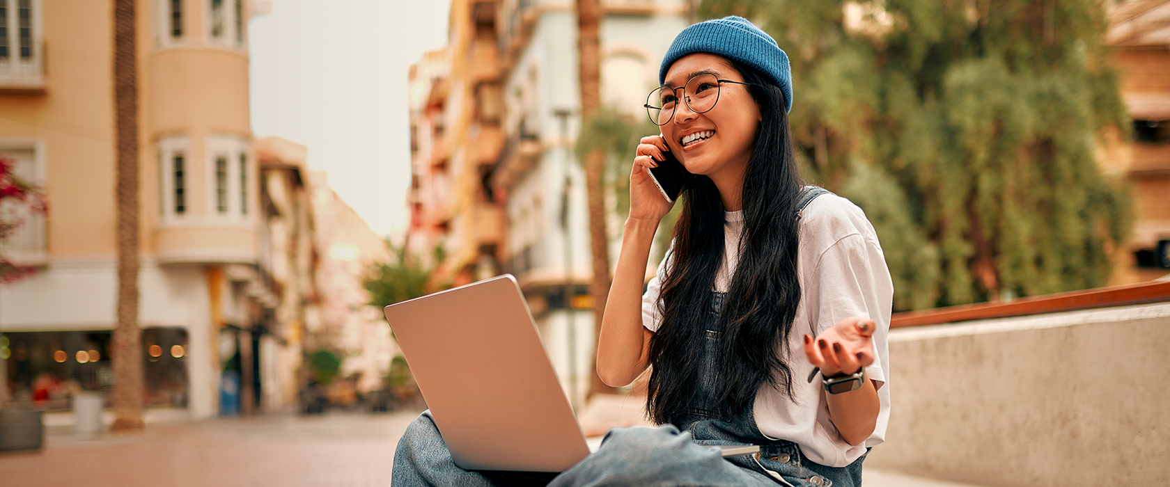 Eine junge Frau mit Brille sitzt im Schneidersitz in einer Promenade. Sie telefoniert, lacht und hat einen Laptop dabei.