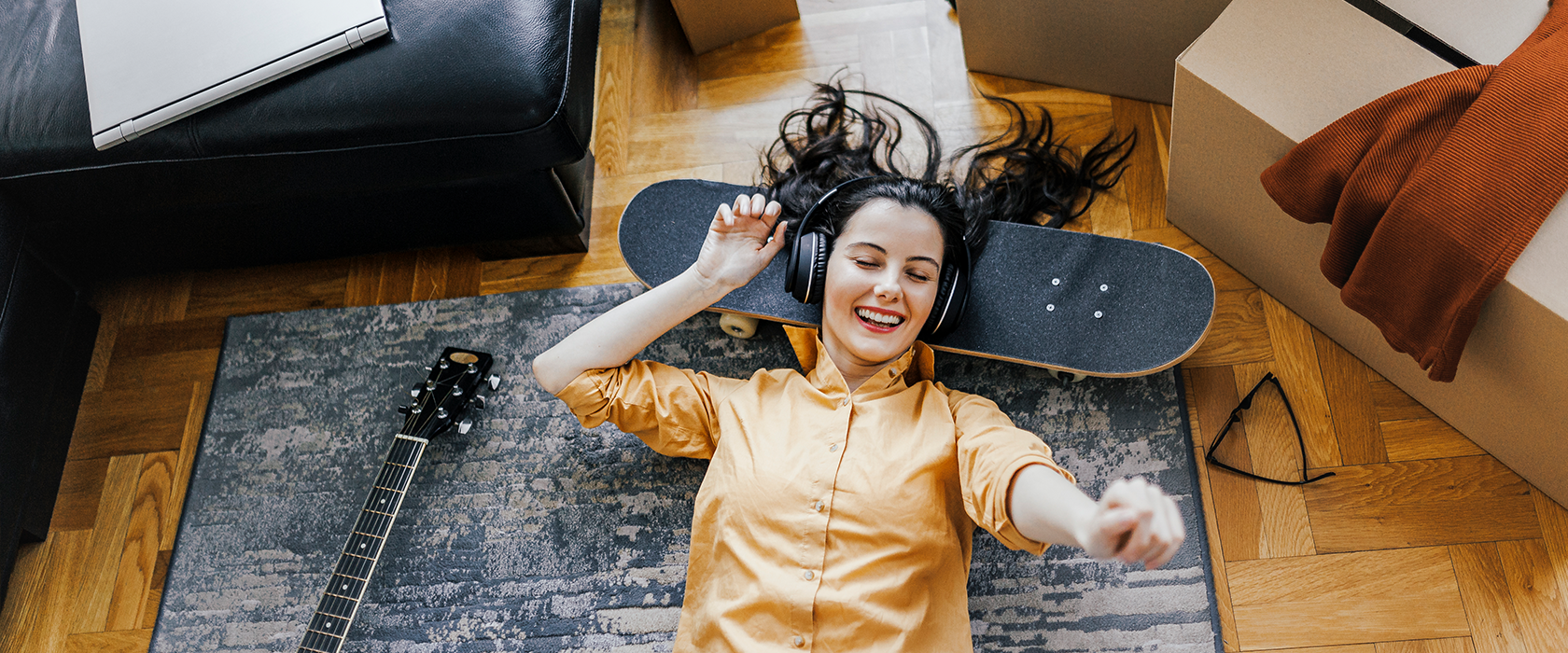 Eine junge Frau chillt zwichen Umzugskartons auf dem Boden und hört Musik. Ein Skateboard dient ihr als Kissen.