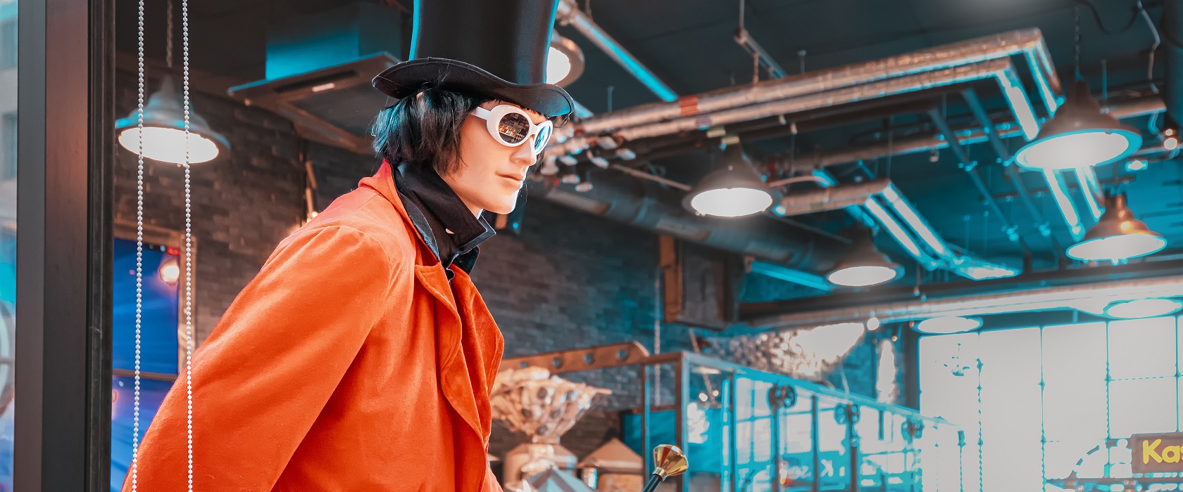 ein als Willy Wonka verkleidetes Mannequin in einer Schokoladenfabrik: es trägt einen Zylinder, einen roten Mantel sowie eine futuristische Sonnenbrille und hat schulterlange, schwarze Haare