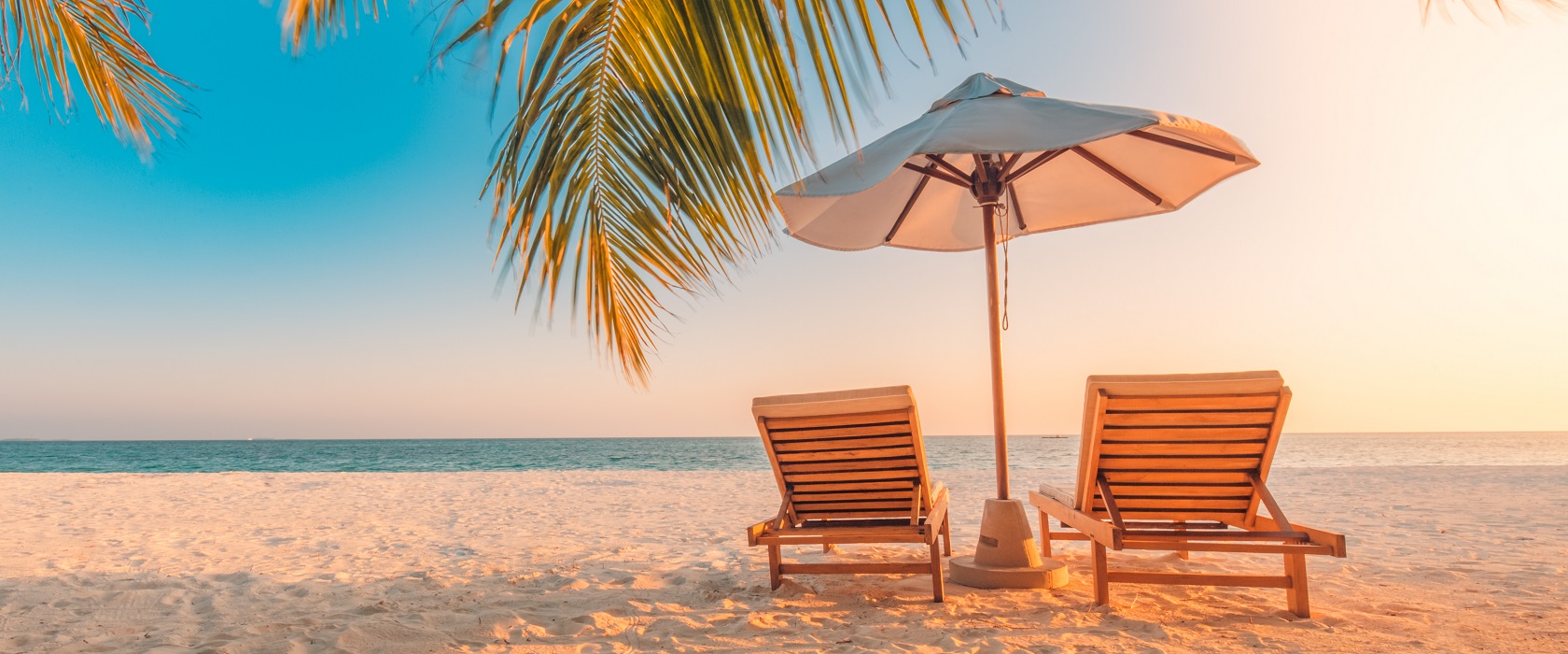 Zwei hölzerne Liegestühle mit einem Sonnenschirm zwischen ihnen, am Strand. Hinter ihnen erstreckt sich türkisblaues Wasser und neben ihnen lugt eine Palme ins Bild. 