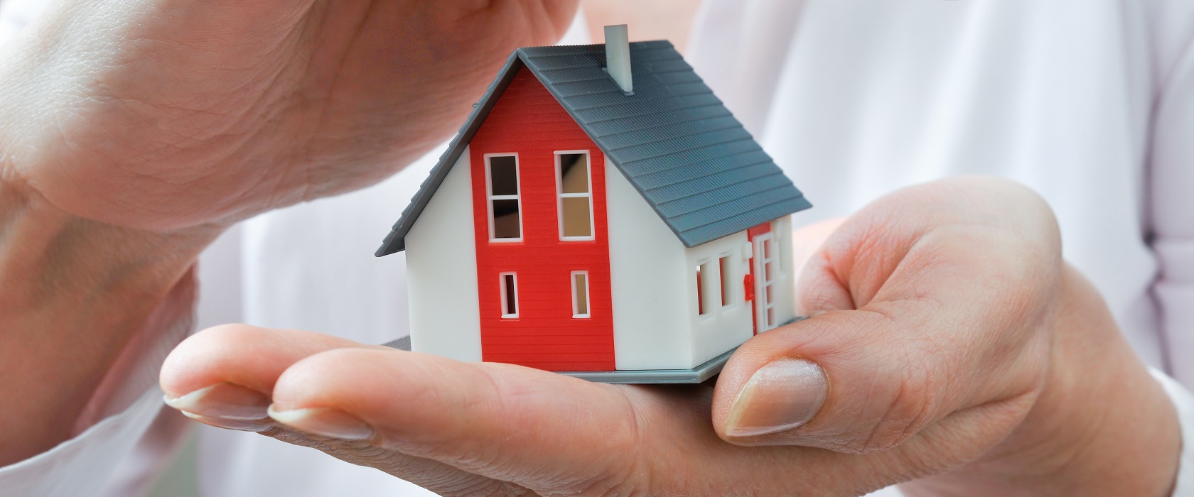 Eine Miniatur-Figur eines Hauses, mit einem rot-weißen Anstrich und einem grauen Giebel, wird in der linken Hand eines Mannes gehalten, während die rechte sich schützend darüberlegt.