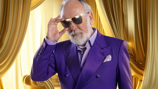 Mann in lila Anzug mit Sonnenbrille, posiert vor goldenen Vorhängen in elegantem Raum.