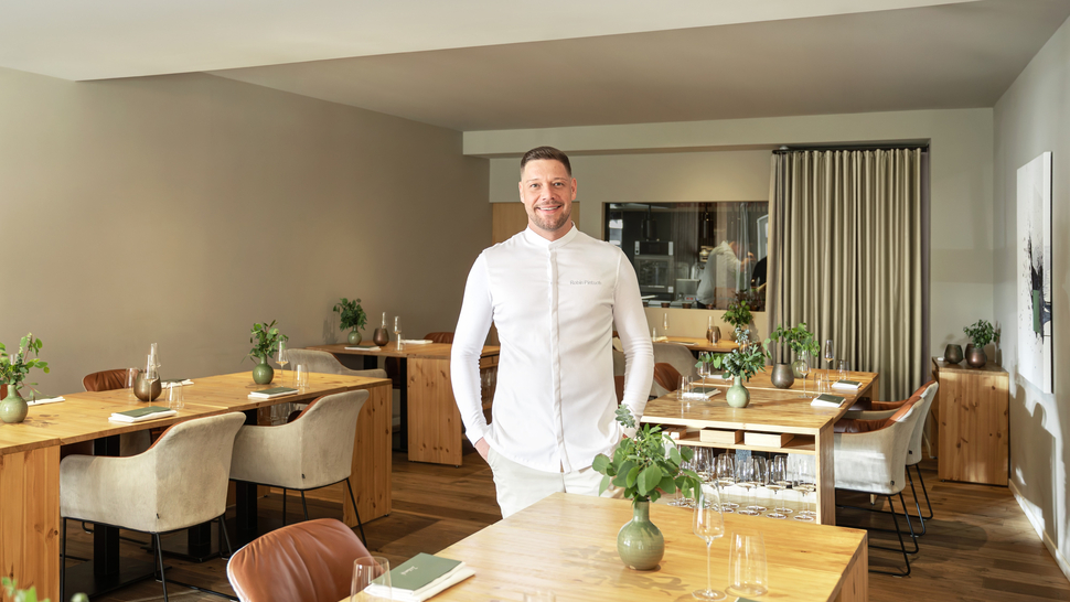 Sternekoch Robin Pietsch in weißem Outfit steht in einem modernen Restaurant mit Holz-Tischen und stilvoller Einrichtung.