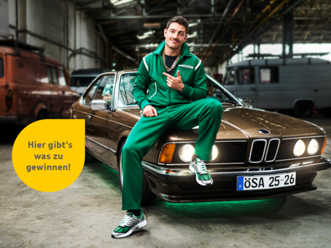 Mann in grünem Trainingsanzug sitzt auf der Motorhaube eines braunen BMWs. Text: „Hier gibt's was zu gewinnen!“