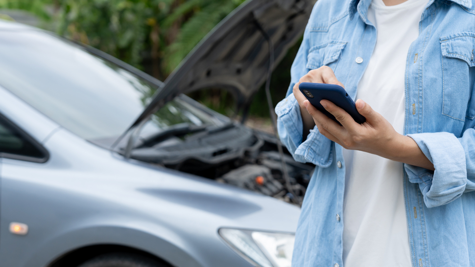 Frau mit Handy vor einem Auto mit offenem Motorhaube, möglicherweise in einer Panne oder auf der Suche nach Hilfe.