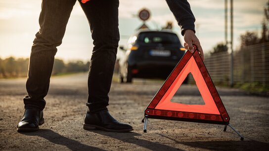 Eine Person platziert ein rotes Warndreieck auf einer Straße, während ein Auto im Hintergrund steht.