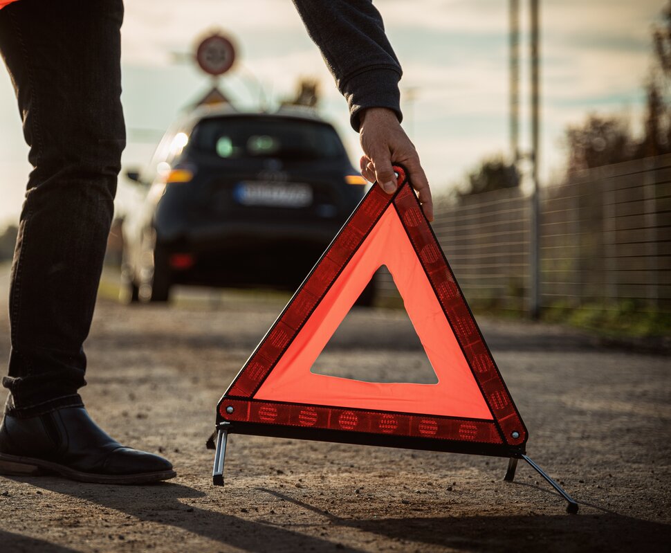 Eine Person platziert ein rotes Warndreieck auf einer Straße, während ein Auto im Hintergrund steht.
