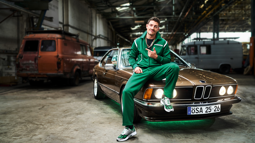Mann in grünem Sportanzug sitzt lässig auf einem braunen BMW in einer Industriehalle. Angedeutetes Lächeln und Daumen hoch.