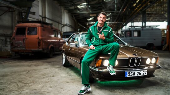Mann in grünem Sportanzug sitzt lässig auf einem braunen BMW in einer Industriehalle. Angedeutetes Lächeln und Daumen hoch.