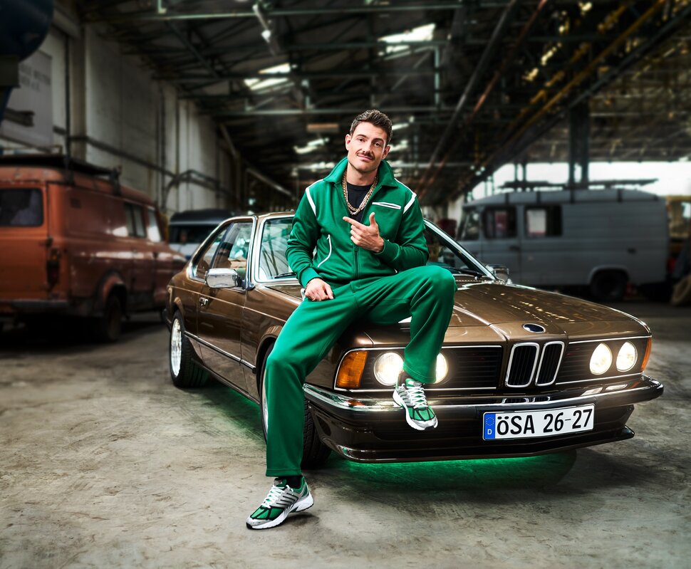 Mann in grünem Sportanzug sitzt lässig auf einem braunen BMW in einer Industriehalle. Angedeutetes Lächeln und Daumen hoch.