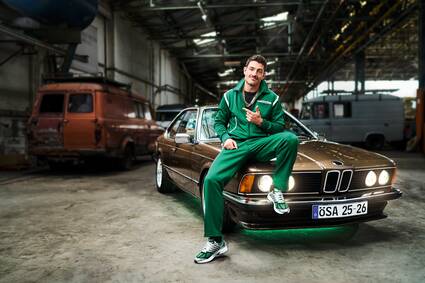 Mann in grünem Sportanzug sitzt lässig auf einem braunen BMW in einer Industriehalle. Angedeutetes Lächeln und Daumen hoch.