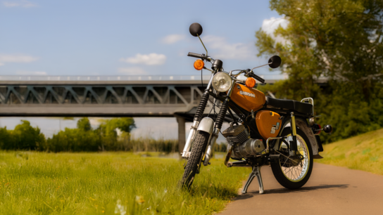 Grauer Roller steht auf einem Feldweg, umgeben von Feldern und unter bewölktem Himmel.