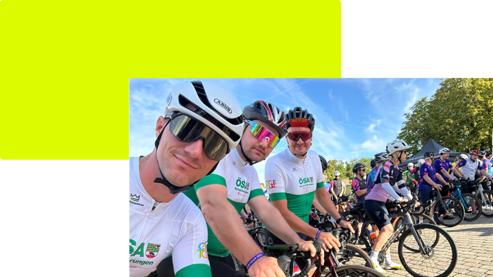Drei Radfahrer in Wettkampfkleidung posieren in einer Gruppe, während weitere Teilnehmer im Hintergrund auf Fahrrädern warten.
