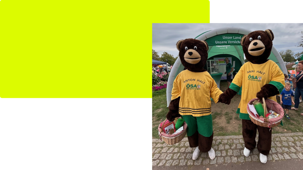 Zwei Bärenkostüme mit gelben T-Shirts stehen nebeneinander und halten Körbe, im Hintergrund ein Zeltdach und viele Besucher.