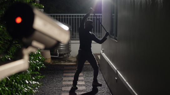 Ein maskierter Einbrecher steht mit einem Gegenstand in der Hand vor einem Haus, das von einer Überwachungskamera beobachtet wird.