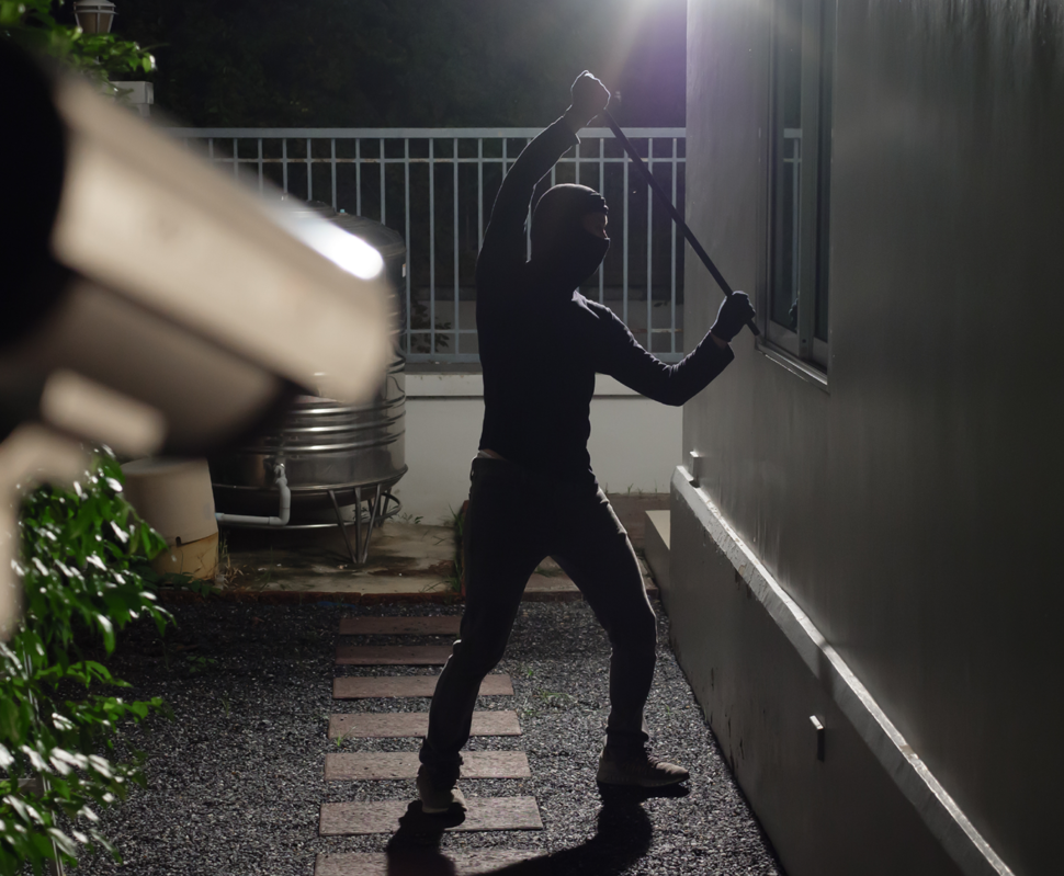Ein maskierter Einbrecher steht mit einem Gegenstand in der Hand vor einem Haus, das von einer Überwachungskamera beobachtet wird.