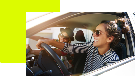 Zwei junge Frauen in einem Auto, eine mit Sonnenbrille am Steuer, lachen und genießen die Fahrt.