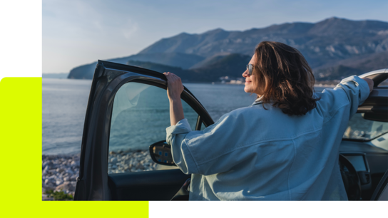 Frau lehnt sich aus dem Autofenster und genießt den Blick auf das Meer und die Berglandschaft im Hintergrund.