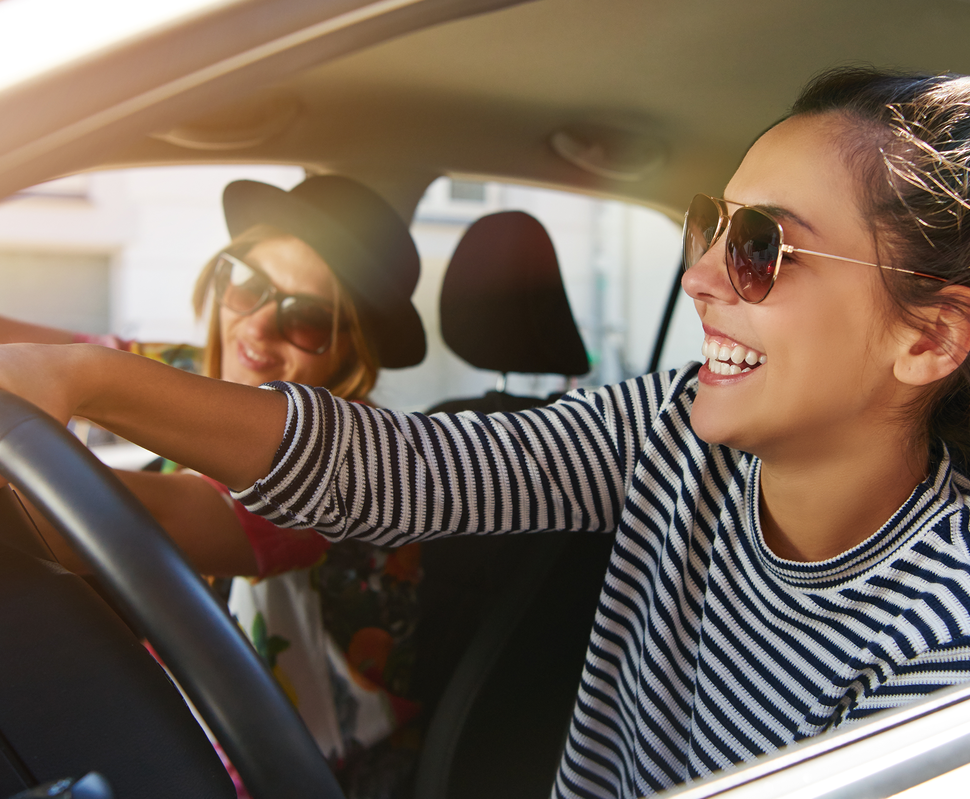 Zwei Frauen im Auto, eine lenkt mit Freude, die andere posiert lächelnd im Hintergrund, beide tragen Sonnenbrillen.