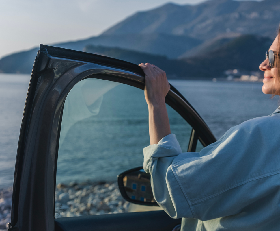 Frau mit Sonnenbrille steht am offenen Autofenster und blickt auf das Meer und die umliegenden Berge.