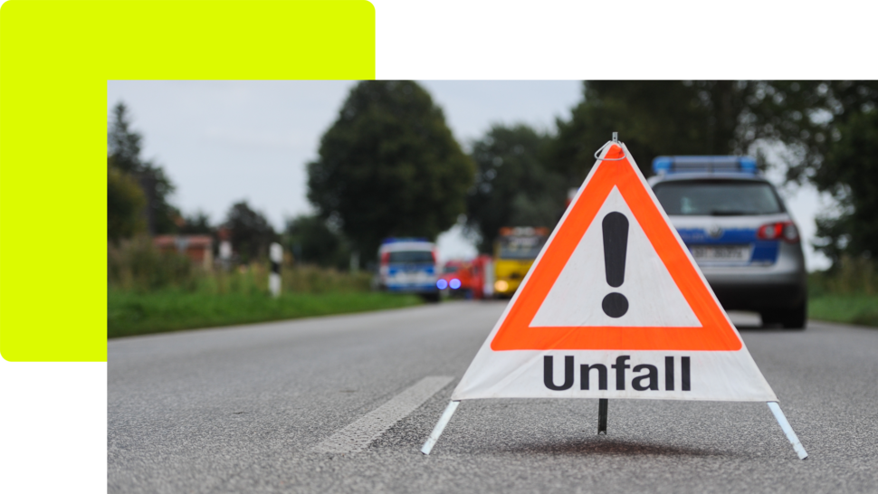 Warnschild mit der Aufschrift „Unfall“ auf einer Straße, im Hintergrund Einsatzfahrzeuge und grüne Landschaft.