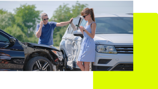 Zwei Personen stehen neben zwei beschädigten Autos auf einem Parkplatz; eine spricht am Telefon, die andere tippt auf ihrem Smartphone.