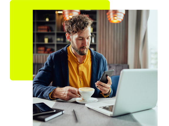 Mann mit lockigen Haaren sitzt an einem Tisch, trinkt Kaffee und schaut auf sein Smartphone, während ein Laptop vor ihm steht.