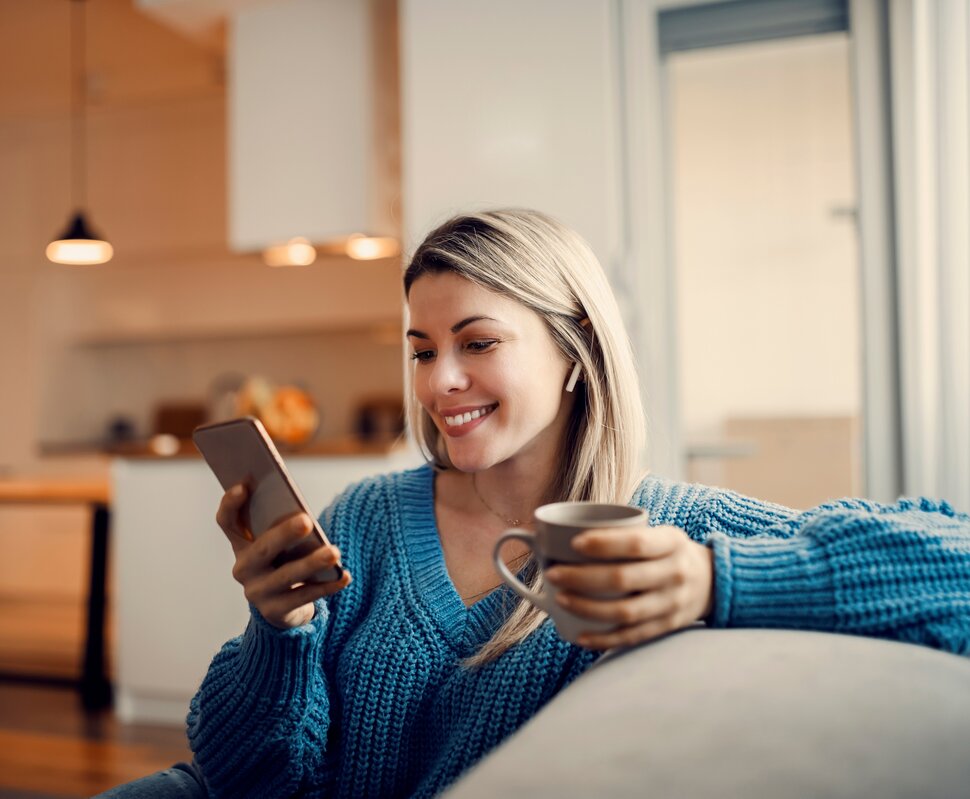 Junge Frau sitzt entspannt auf einer Couch, hält ein Smartphone und lächelt, während sie es nutzt.