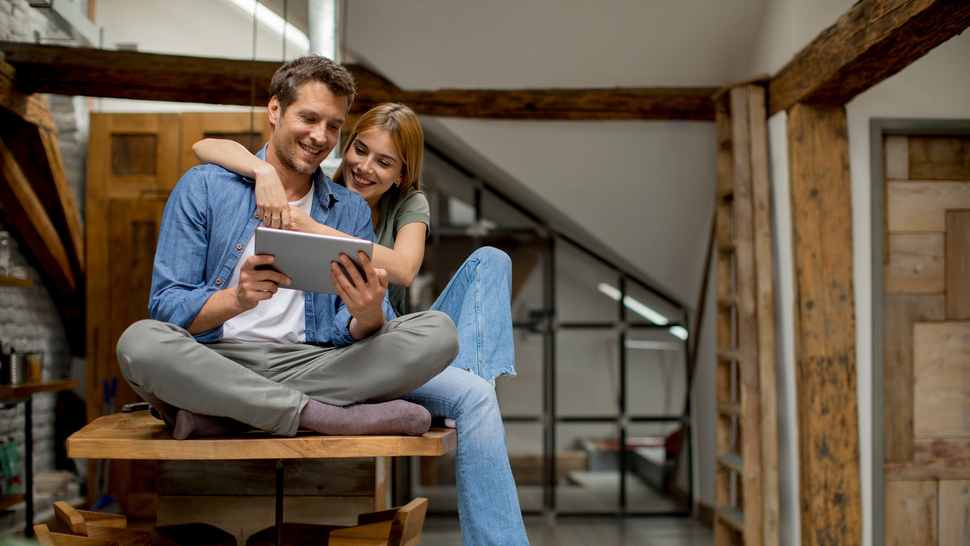 Junges Paar sitzt auf einem Tisch, lächelt und schaut zusammen auf ein Tablet in einer modern eingerichteten Wohnung.
