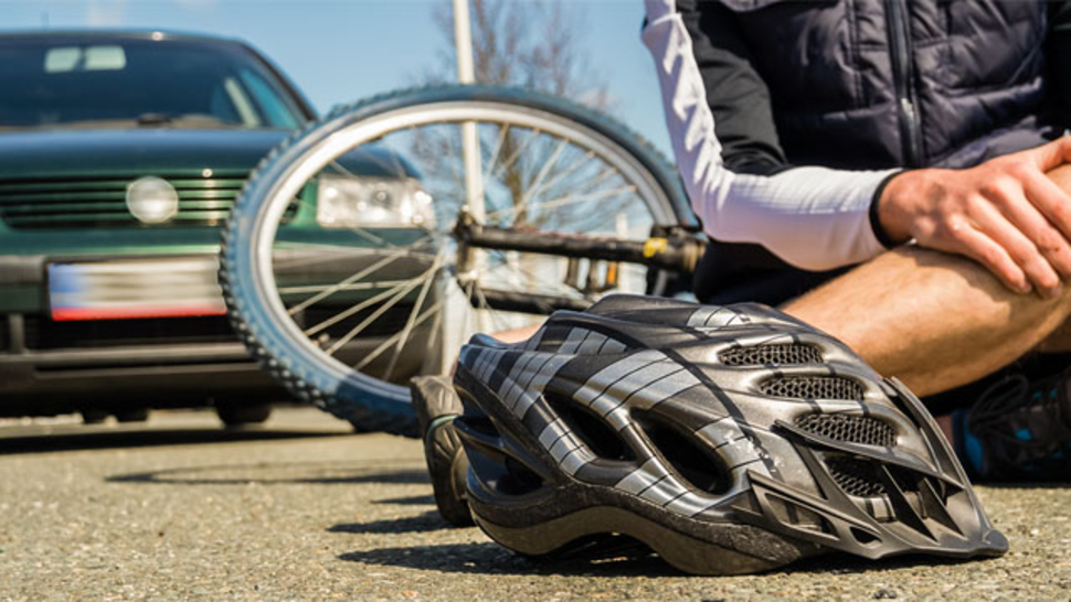 Fahrradhelm liegt auf dem Boden, während ein Radfahrer neben seinem Fahrrad sitzt, nahe einem geparkten Auto.