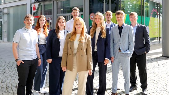 Gruppe von jungen Menschen in modernen Outfits, die in einem urbanen Umfeld auf einem Platz stehen und lächeln.