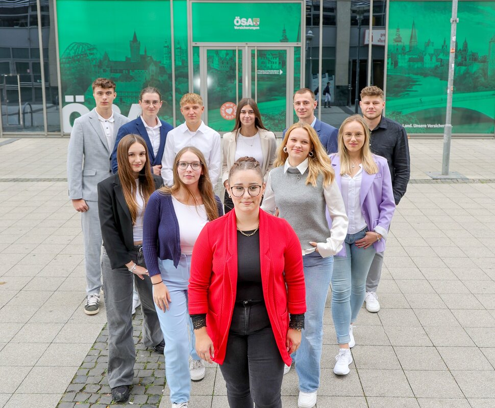 Gruppe von jungen Menschen in modernen Outfits, die in einem urbanen Umfeld auf einem Platz stehen und lächeln.