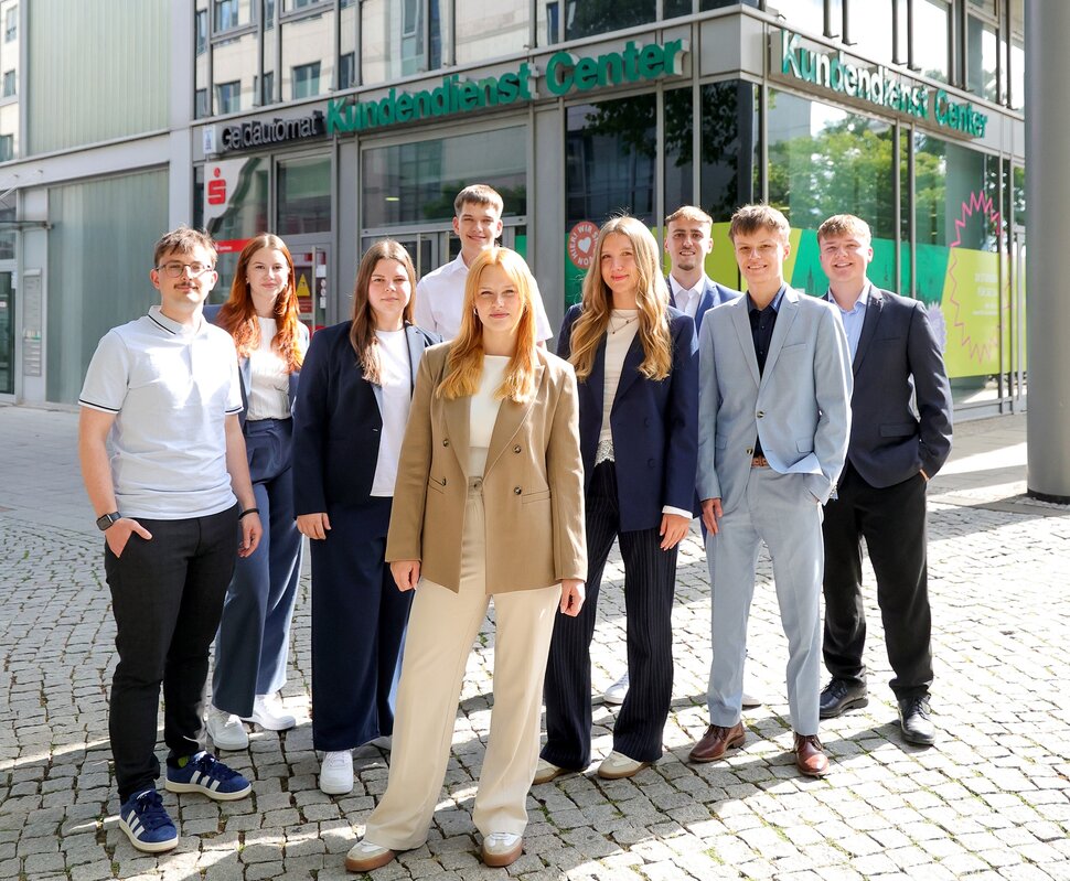 Gruppe von jungen Menschen in modernen Outfits, die in einem urbanen Umfeld auf einem Platz stehen und lächeln.