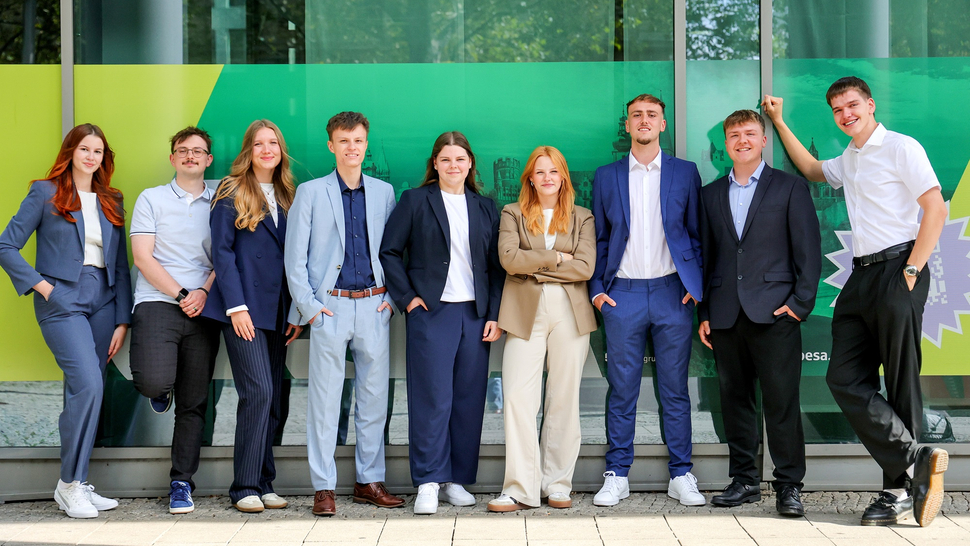 Gruppe junger Erwachsener in formeller Kleidung posiert vor einer bunten Stadtszene. Fröhliche Gesichter, freundliche Stimmung.