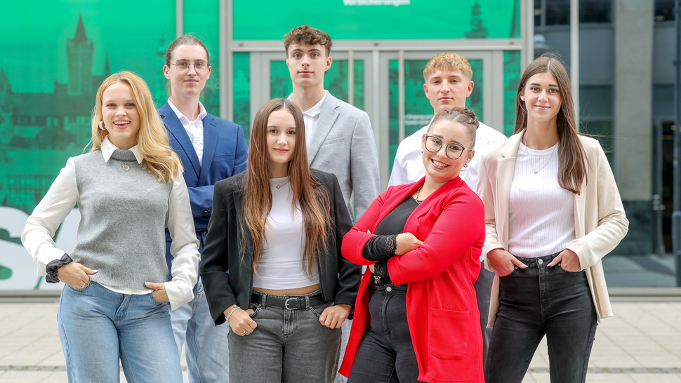 Gruppe junger Erwachsener in formeller Kleidung posiert vor einer bunten Stadtszene. Fröhliche Gesichter, freundliche Stimmung.