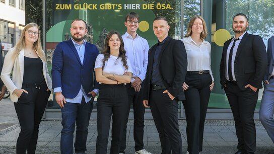 Neun Personen in formeller Kleidung stehen vor einem modernen Gebäude mit buntem Schriftzug im Hintergrund.