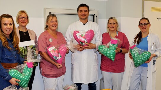 Sechs Personen in Krankenhauskleidung stehen lächelnd zusammen und halten Geschenke in Form von Herzen.