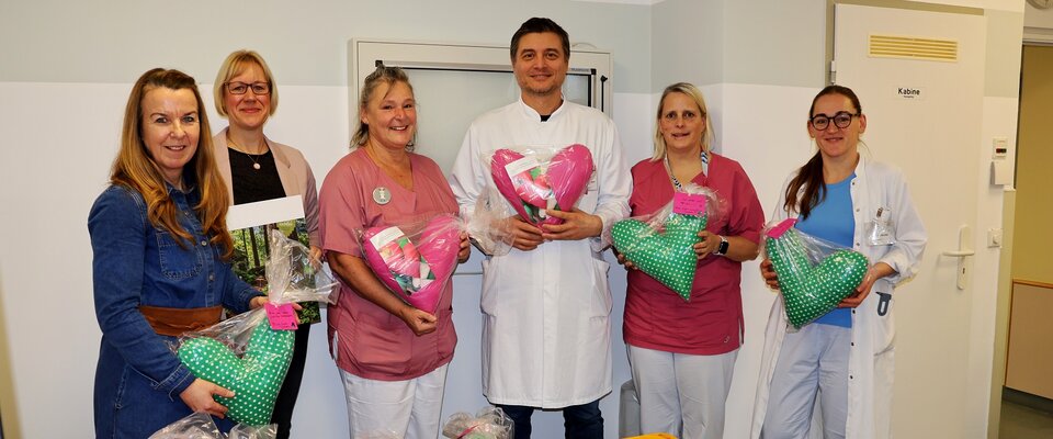 Sechs Personen in Krankenhauskleidung stehen lächelnd zusammen und halten Geschenke in Form von Herzen.
