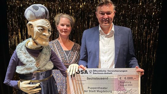 Eine Puppenspielerin und ein Mann halten einen Scheck über 6.000 Euro für das Puppentheater der Stadt Magdeburg.