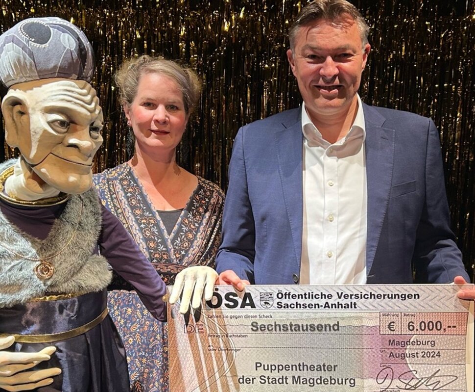 Eine Puppenspielerin und ein Mann halten einen Scheck über 6.000 Euro für das Puppentheater der Stadt Magdeburg.