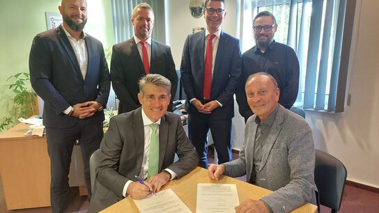 Vorstand David Bartusch und Bürgermeister Jörg Methner unterzeichnen den Feuerwehr-Renten-Rahmenvertrag, umgeben von vier Personen im Hintergrund in formeller Kleidung.