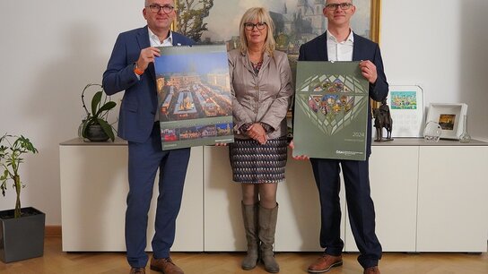Drei Personen posieren in einem Büro und halten Plakate mit Stadtansichten und einem Kalender für 2023.