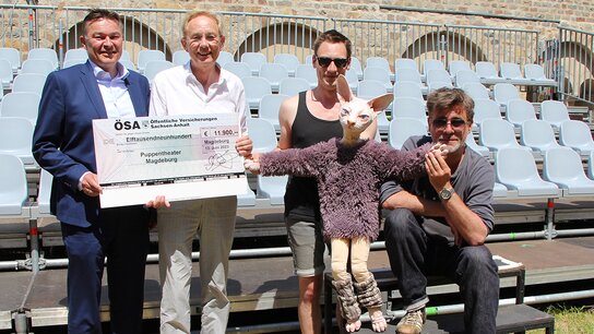 Vier Personen halten einen großen Scheck und posieren auf einer Tribüne; eine trägt ein Haustier in einem Pullover.